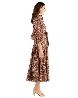 A woman modeling a long-sleeved midi dress with a detailed multicolor paisley and floral pattern in red, green, and beige, featuring a tie waist and flared cuffs, shown from a side profile against a light gray studio background.