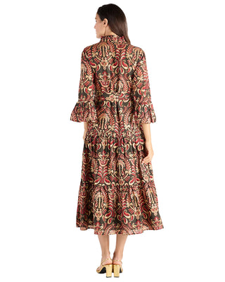 A woman viewed from the back wearing a long-sleeve midi dress with a dense botanical and paisley-style print in rust red, beige, and black, paired with gold heeled sandals against a plain white studio background.