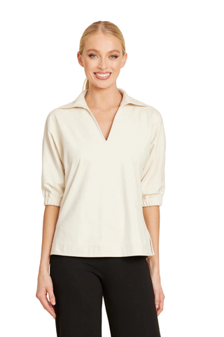 A woman modeling a cream-colored, three-quarter sleeve collared top with a V-neckline, paired with black pants, against a solid light beige background.