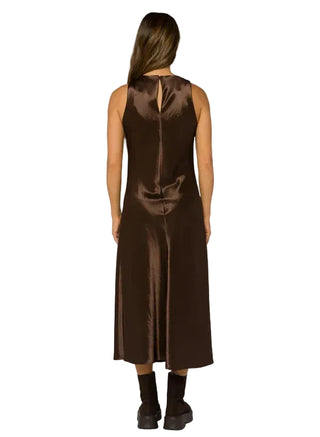 A woman viewed from the back wearing a sleeveless, knee-length brown satin dress with a back slit and a small keyhole cutout at the top, paired with dark brown ankle boots, against a light gray studio background.