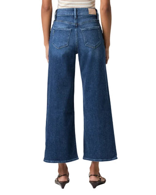 A model shown from the back wearing a pair of wide-leg cropped blue denim jeans, paired with a striped top and black sandals, against a plain light gray studio background.