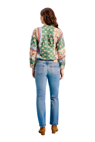 A woman is shown from behind wearing a green bohemian-style patterned blouse and blue jeans, standing against a solid light blue background.