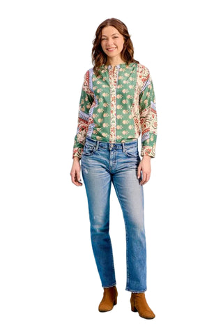 A woman modeling a green bohemian-style patterned blouse with colorful paisley sleeves, paired with blue jeans and brown suede ankle boots, against a solid light blue studio background.