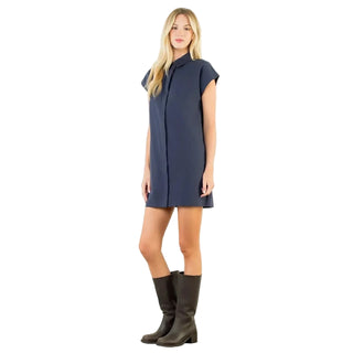 A woman modeling a navy blue short-sleeve collared shirt dress with dark brown knee-high boots, standing against a plain white studio wall.