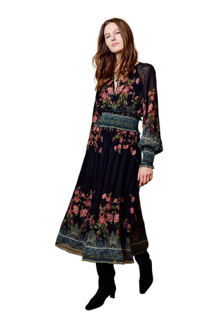 A woman models a long-sleeved midi dress with a dark navy base and a pink and green floral pattern, paired with black boots, against a solid tan studio backdrop.