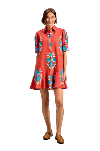 A woman modeling a short-sleeve collared dress with a coral-red base and blue, white, and yellow abstract floral patterns, paired with brown loafers, against a solid light blue background.