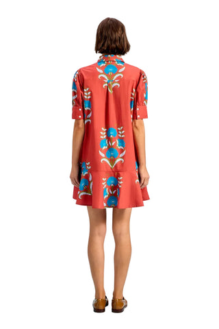 A woman viewed from the back wearing a coral-red short-sleeve shirt dress with a blue, beige, and white geometric pattern, set against a solid light blue background.
