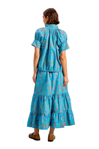 A woman shown from the back wearing a matching short-sleeve blouse and tiered midi skirt set in bright turquoise blue with a pink paisley pattern, paired with brown flat shoes, against a solid light blue studio background.