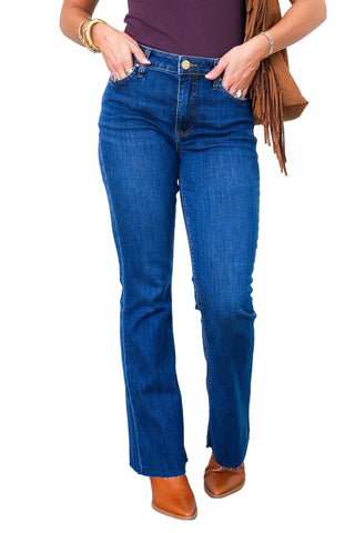 A person wearing dark blue bootcut jeans, styled with a brown fringe bag and brown leather boots, standing against a white paneled wall.