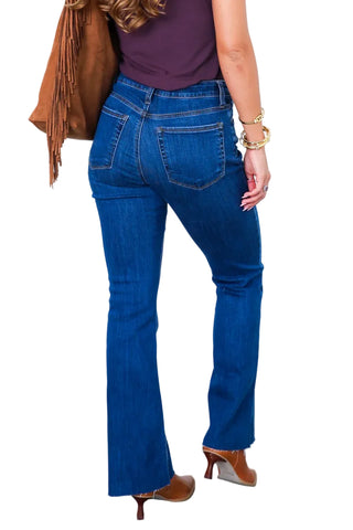 A person shown from the back wearing blue flare jeans, a dark purple sleeveless top, brown fringe shoes, and a brown shoulder bag against a white paneled wall and gray floor.