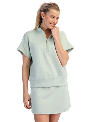 A woman modeling a light sage green two-piece lounge set consisting of a short-sleeve half-zip pullover and a matching mini skirt, against a light gray studio background.