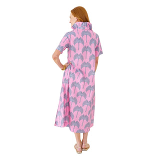 A woman stands with her back to the camera wearing a pink dress with a gray palm tree pattern, set against a white brick wall background.