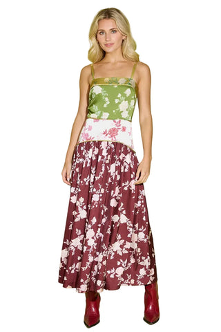 A woman modeling a sleeveless maxi dress with a green floral bodice, a white waistband with pink floral accents, and a burgundy floral skirt, paired with burgundy boots, against a light green studio background.