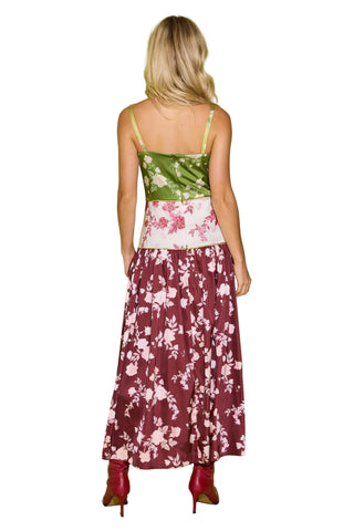 A woman stands with her back to the camera wearing a sleeveless maxi dress with a green satin bodice, a white waistband with pink floral print, and a long burgundy skirt with a pink floral pattern. She is wearing burgundy pointed-toe heels against a solid light green studio background.