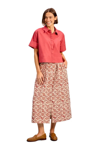 A woman modeling a coral pink short-sleeve button-up shirt and a high-waisted, wide-leg midi skirt with a red and beige floral block print, paired with brown leather loafers, against a solid light blue studio background.