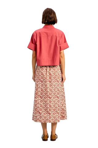 A model viewed from the back wearing a coral pink short-sleeve button-up shirt and a pink patterned midi skirt, set against a solid light blue studio background.
