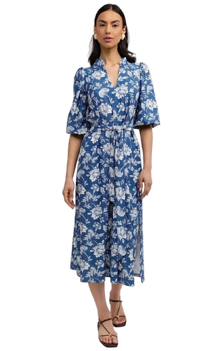A woman modeling a blue and white floral midi wrap dress with puff sleeves and a tie waist, paired with brown strappy sandals, against a light gray studio background.