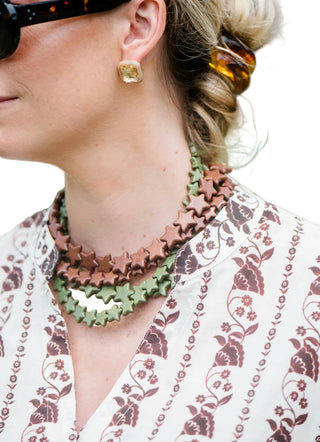 A close-up lifestyle shot of a woman wearing a layered necklace with star-shaped links in terracotta brown and sage green, paired with a cream blouse featuring a brown floral pattern.