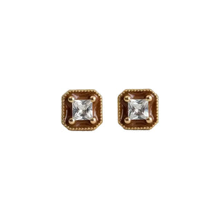 A pair of gold square stud earrings with a central clear princess-cut gemstone and a textured gold border, displayed on a light gray textured fabric background.