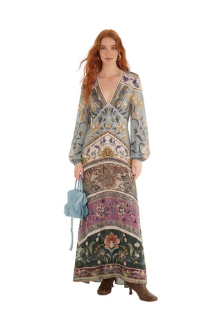 A model with red hair stands against a plain white studio background wearing a long-sleeved maxi dress with an intricate, multi-colored ornamental pattern. She is holding a small light blue handbag and wearing brown heeled boots.