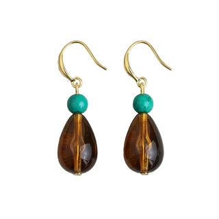 A pair of handmade dangle earrings featuring teardrop-shaped amber brown beads, a small round turquoise bead, and gold-tone fishhook wires, displayed on a textured light gray fabric background.
