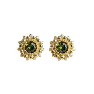 A pair of gold flower-shaped stud earrings with round green crystal centers, displayed on a light gray textured fabric background.