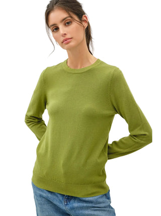 A woman wearing a light olive green crewneck sweater and blue jeans, photographed against a neutral light gray studio background.