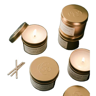 Several Charleston Candle Co. soy candles in round gold metal tins are arranged on a dark green surface, with two lit candles, one open candle with its lid beside it, and a few wooden matches.